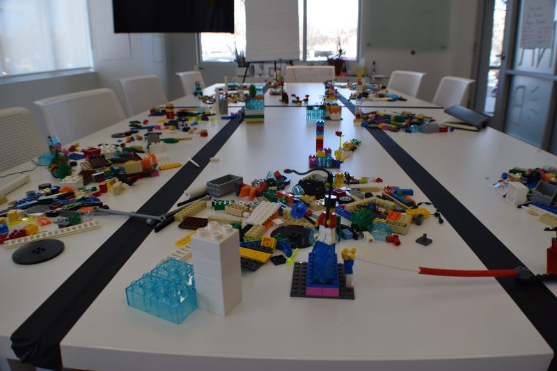 A long conference table covered in models and loose bricks, chairs pulled back, mid-session